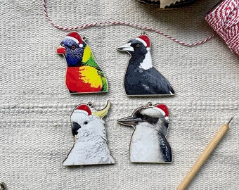 May include: Four wooden bird ornaments, each wearing a Santa hat. The birds include a rainbow lorikeet, a black and white magpie, a white cockatoo, and a kookaburra. The ornaments are on a textured, light-colored surface.