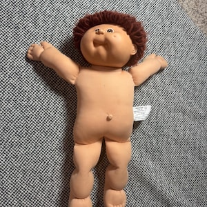 May include: A Cabbage Patch Kids doll with a soft peach-colored fabric body, a vinyl head, and short brown yarn hair. The doll has a smiling expression, brown eyes, and a small belly button. A white fabric tag is attached to the side of the torso.