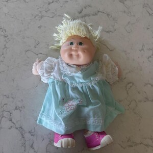 Boneca Cabbage Patch Kids com cabelo loiro de lã, olhos verdes, vestido verde menta e sapatos rosa.
