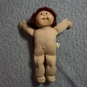 Op de afbeelding: Een Cabbage Patch Kids pop met een zacht, lichtgekleurd stoffen lichaam, een rond vinyl hoofd en kort, krullend bruin garenhaar. De pop heeft een kleine navel, uitgestrekte armen en een label aan de zijkant, met een glimlachende gezichtsuitdrukking.