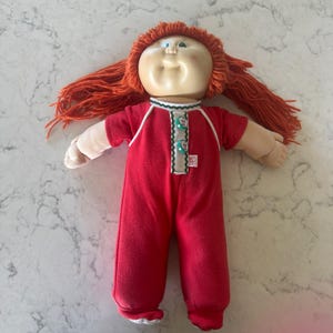 Könnte beinhalten: Eine Cabbage Patch Kids Puppe mit leuchtend orangefarbenem Wollhaar und rundem Gesicht. Die Puppe trägt einen roten Einteiler mit weißem Saum, einer Knopfleiste mit zwei türkisfarbenen Knöpfen und einem kleinen weißen Etikett mit CPK.