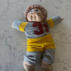 Puede incluir: Una muñeca de cuerpo blando con cabello de lana marrón claro y ojos azules, que viste una camiseta gris y amarilla con el número 31 impreso en rojo en el pecho, junto con pantalones cortos a juego. Tiene cabeza de vinilo y extremidades de tela.