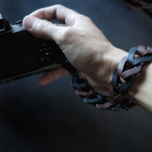 Può includere: Un cinturino da polso per fotocamera realizzato con anelli intrecciati di pelle testurizzata nera e marrone. Il cinturino presenta un design intrecciato che si fissa attorno al polso, con un'estremità collegata al corpo di una fotocamera.