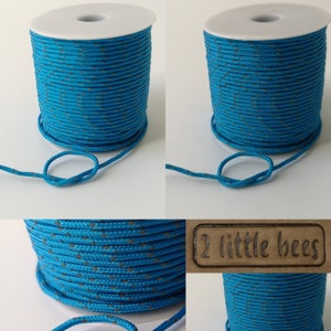 May include: A spool of turquoise blue cord with a grey diamond pattern. The cord is wound around a white plastic spool. The spool is labeled "2 little bees".