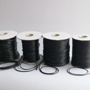 May include: Four spools of black leather cord, each wound around a white cardboard spool. The spools are arranged in a row on a white surface.