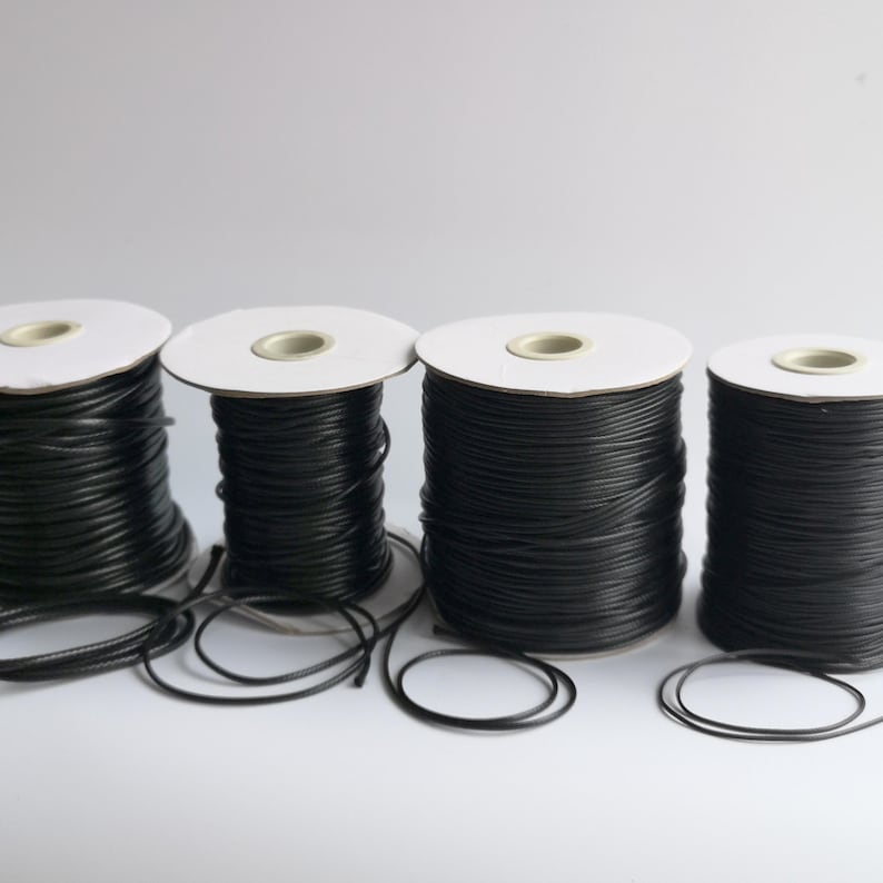 May include: Four spools of black leather cord, each wound around a white cardboard spool. The spools are arranged in a row on a white surface.