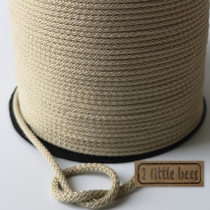 May include: A spool of beige cotton rope with a black edge. The rope is coiled neatly and has a small loop at the end. A brown label with the text "2 little bees" is attached to the rope.