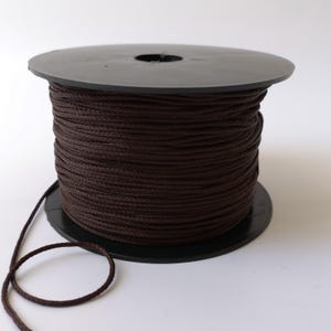 May include: A spool of brown waxed cotton cord. The cord is wound tightly around a black plastic spool. A length of cord is hanging off the spool.