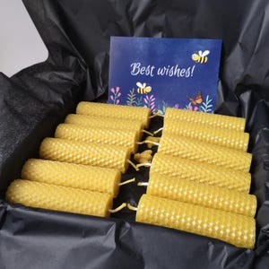 May include: A gift box filled with yellow honeycomb-patterned candles. A blue card with the words "Best wishes!" and illustrations of bees and flowers is also in the box. The candles are cylindrical and have a natural, warm color.