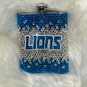 Puede incluir: Una petaca rectangular de acero inoxidable cubierta de pedrería azul y plateada. La palabra "LIONS" está escrita en letras blancas en el centro. La petaca tiene una tapa y una bisagra plateadas.