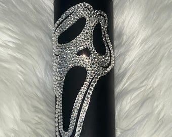 May include: Black tumbler featuring a Scream mask design, embellished with sparkling rhinestones. The mask is outlined with rhinestones, contrasting against the matte black surface. The tumbler has a black lid.