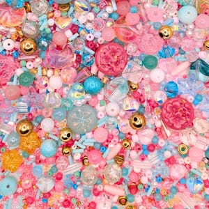 May include: A collection of assorted craft beads in various shapes, sizes, and textures. The mix includes pink, blue, clear, and gold-toned beads, featuring floral patterns, smooth spheres, faceted crystals, and small cylindrical seed beads.