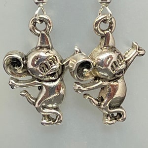 May include: A pair of silver-toned earrings featuring cartoon-like figures. Each earring has a small, detailed figure with large ears, arms outstretched, and a tail. The earrings hang from a simple silver hook.