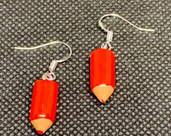 May include: A pair of novelty earrings shaped like red pencils with silver-colored hooks. The pencil earrings have a bright red body and a tan-colored tip, resembling a sharpened pencil. The earrings are displayed against a textured, dark gray background.
