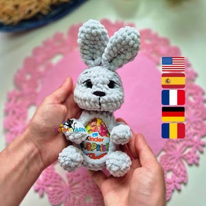 Könnte beinhalten: Ein hellgrauer gehäkelter Hase mit großen Ohren und schwarzen Augen. Der Hase hält ein Kinder Überraschungsei. Im Hintergrund befinden sich eine rosa Tischdecke und ein dekoratives Osterei.