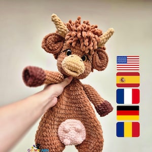 Könnte beinhalten: Ein handgefertigtes Highland-Kuh-Plüschtier aus braunem und beigem Häkelgarn. Das Plüschtier hat ein hellbraunes Gesicht, Ohren und Hörner, mit einem dunkelbraunen Körper und Beinen. Die Kuh hat einen rosa Bauch.