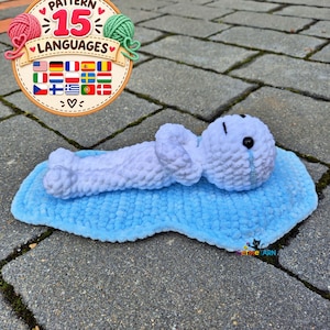 May include: A white crocheted plush figure lying on a blue crocheted puddle. The figure has black bead eyes and a blue tear stitched under one eye. A circular graphic in the upper left corner displays the text Pattern 15 Languages with yarn icons.
