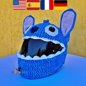 May include: A blue crochet helmet cover featuring a character design with large upright ears, white eye patches with black pupils, and a dark blue nose. The cover has a scalloped white border around the dark tinted visor opening and a logo.
