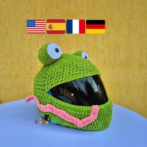 Può includere: Una copertura per casco all'uncinetto di colore verde brillante con due grandi occhi bianchi e neri sulla parte superiore e una lunga lingua rosa che si estende sul davanti. La copertura è progettata per un casco da moto con visiera scura.