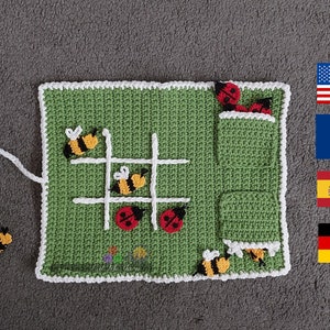 Può includere: Un gioco di tris in crochet verde con un bordo bianco. Il tabellone di gioco ha una corda bianca attaccata ad esso. Ci sono coccinelle rosse e api gialle e nere sul tabellone. Ci sono anche due tasche sul lato del tabellone. Le tasche sono piene di altre coccinelle e api. Il gioco è su una superficie grigia. Ci sono quattro bandiere sullo sfondo: americana, francese, spagnola e tedesca.