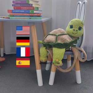 May include: A crocheted green turtle backpack with a brown shell and a smiling face. The backpack is sitting on a wooden chair with bunny ears. The chair is in front of a white curtain and a table with a stack of books and flags of the United States, Germany, France, and Spain.