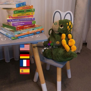 Może przedstawiać: Zielono-żółta pluszowa zabawka w kształcie dinozaura siedząca na szarym drewnianym krześle. Krzesło ma białą, pomalowaną podstawę. Zabawka siedzi przed małym drewnianym stolikiem z szarym blatem. Na stoliku leży stos książek dla dzieci. Książki mają tytuły w języku rumuńskim i angielskim. Na stoliku znajdują się flagi Stanów Zjednoczonych, Francji i Hiszpanii.