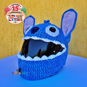 Puede incluir: Una funda de ganchillo azul para casco diseñada para parecerse a un personaje con orejas grandes y erguidas, una nariz oscura y parches blancos en los ojos. La funda presenta un borde blanco dentado alrededor de la visera oscura e incluye un logotipo circular con el texto Pattern 15 Languages y GetMeYarn.