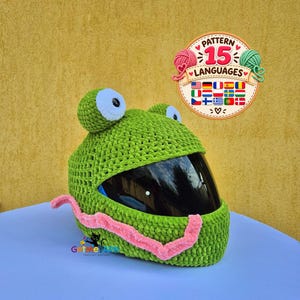 Puede incluir: Una funda de ganchillo verde para casco con forma de rana, con grandes ojos blancos y negros en la parte superior y una larga lengua rosa en la parte delantera. Una insignia circular en la esquina superior derecha muestra el texto Pattern 15 Languages.