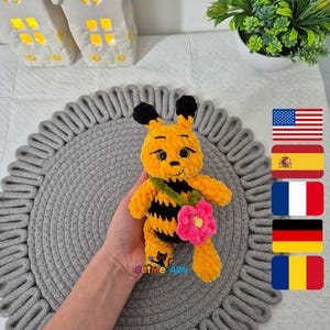 May include: A crocheted bee plush toy with black and yellow stripes, holding a pink flower. The bee has a friendly face and is sitting on a gray crocheted mat. The image also includes flags of the United States, Spain, France, Germany, and Romania.