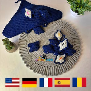 May include: A blue crocheted shark blanket with a set of smaller crocheted shark toys. The toys are arranged on a gray rope rug. The image also includes flags of the United States, Germany, France, Spain, and Romania.