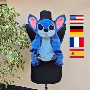 Może przedstawiać: Niebieska i jasnoniebieska szydełkowa pluszowa zabawka przedstawiająca postać z kreskówki z dużymi uszami i czarnymi oczami. Plusz jest wystawiony na czarnym manekinie. Tekst "get me YARN" jest widoczny na pluszaku. Flagi USA, Niemiec, Francji i Hiszpanii w tle.