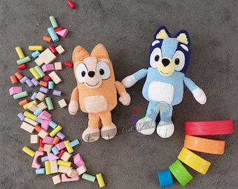 May include: Two crocheted stuffed animals, one orange and one blue, with large eyes and smiling faces. The orange animal is a cat and the blue animal is a dog. The animals are sitting on a gray surface with colorful wooden blocks scattered around them.