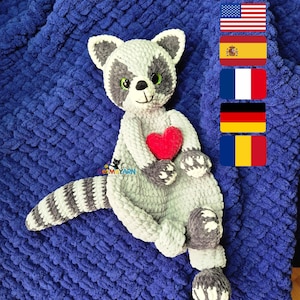 Puede incluir: Un peluche artesanal de mapache en tonos grises, blancos y negros, sosteniendo un corazón rojo. El mapache está sobre una manta azul oscuro texturizada. Los ojos del mapache son verdes.