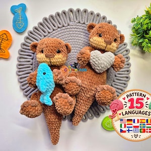 May include: Two brown crocheted otter plush toys made of soft yarn. One otter holds a small bright blue crocheted fish, while the other holds a light gray crocheted heart. A circular badge with the text PATTERN 15 LANGUAGES is visible.
