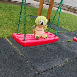 May include: A green and brown crocheted turtle plush toy sits on a red swing set. The swing set is made of plastic and has green ropes.