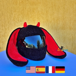 May include: A black crochet helmet cover featuring two small red horns on top and two long, wide, floppy ears with red inner lining. The cover has a dark, reflective visor opening and is designed to fit over a standard motorcycle helmet.