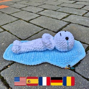 Puede incluir: Una figura de peluche tejida a ganchillo en color blanco con cabeza redonda, ojos de cuentas negras y una lágrima azul bordada, acostada sobre una alfombrilla de ganchillo azul claro con forma de charco. Tiene una textura de hilo suave.
