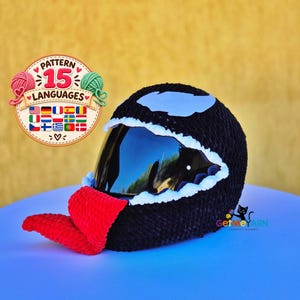 May include: A black crochet helmet cover featuring a large red tongue, white jagged teeth trim around the visor, and white eye patches. A circular badge displays the text Pattern 15 Languages with various flags, and a logo reads GetMeYarn.