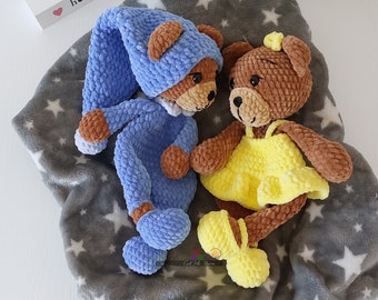 May include: Two crocheted teddy bears wearing pajamas and a dress, lying on a gray blanket with white stars. The teddy bear wearing pajamas is blue and has a sleeping cap. The teddy bear wearing a dress is brown and yellow. The blanket has the text "Get Me Yarn" on it.