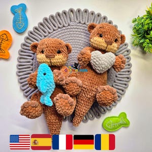 May include: Two brown crocheted otter plush toys, one holding a blue fish and the other a gray heart. The otters are on a gray circular mat, with felt fish decorations in blue, orange, and green.