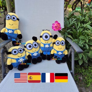 May include: A collection of five crocheted Minion dolls, each with yellow bodies, blue overalls, and black accents. They wear goggles and have a white flower detail. The dolls are displayed on a chair with a pink flower and greenery in the background.