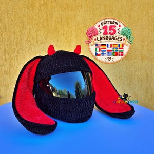May include: A black crochet helmet cover featuring long, red-lined bunny ears and two small red devil horns on top. The cover is designed to fit over a motorcycle helmet with a dark, reflective visor. A circular logo with the text PATTERN 15 LANGUAGES is visible.