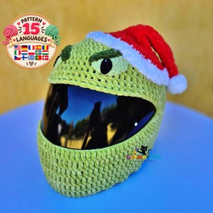 May include: A light green crochet helmet cover featuring a red and white Santa hat on top. The cover has dark green crocheted eyebrows and black oval eyes positioned above a large, dark, reflective visor opening. The texture is thick and yarn-based.