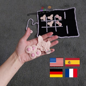 May include: A crocheted tic-tac-toe game with pink and white pieces. The game board is black with white lines and is folded in half. The game is laid out on a gray surface. The image also includes the flags of the United States, Spain, Germany, and France.