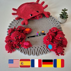 Puede incluir: Un cangrejo rojo de crochet con una gran concha y cangrejos más pequeños dentro. El cangrejo grande está sentado en una alfombra de cuerda gris con cangrejos rojos más pequeños a su alrededor. El texto "Get Me Yarn" es visible en la alfombra. Hay banderas de los Estados Unidos, España, Francia, Alemania y Rumania en la parte inferior de la imagen.