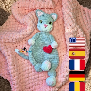 May include: A handmade, light blue cat-shaped baby blanket with a red heart and white accents. The blanket is a soft, pink color and has a woven texture. The cat has green eyes and a pink nose. The blanket measures approximately 30 inches by 30 inches.