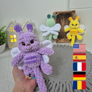 Puede incluir: Un juguete de mariposa de crochet morado con alas blancas. El juguete está sentado en una alfombra gris con un patrón tejido. Hay banderas de Estados Unidos, España, Francia, Alemania y Rumania en el fondo.