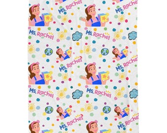 May include: A white blanket featuring a repeating pattern of Ms. Rachel wearing a pink headband and blue overalls, holding a yellow block. The design includes colorful polka dots, yellow suns, blue clouds, globes, and the text Ms. Rachel in pink and blue.