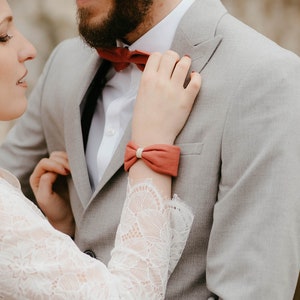 May include: A couple in formal attire. The woman wears a white lace top and skirt, the man a grey suit with a red bow tie. The woman has a matching red bracelet. The background is a stone wall and greenery.