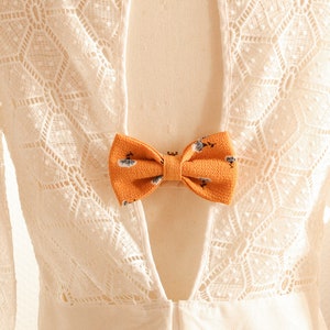 May include: A white lace dress with a low back and a yellow bow tie. The dress is on a mannequin and the background is a white wall with a gold geometric decoration.